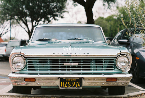 A vintage 1966 Chevrolet Nova in a light teal green color, parked outdoors in a lot with trees in the background. The front of the car is facing the camera, showing a chrome grille, round headlights, and a California license plate.