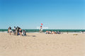 A wide beach scene with sand in the foreground, ocean water in the distance, several small groups of people near the shoreline, and a lifeguard tower with a red flag under a clear blue sky.