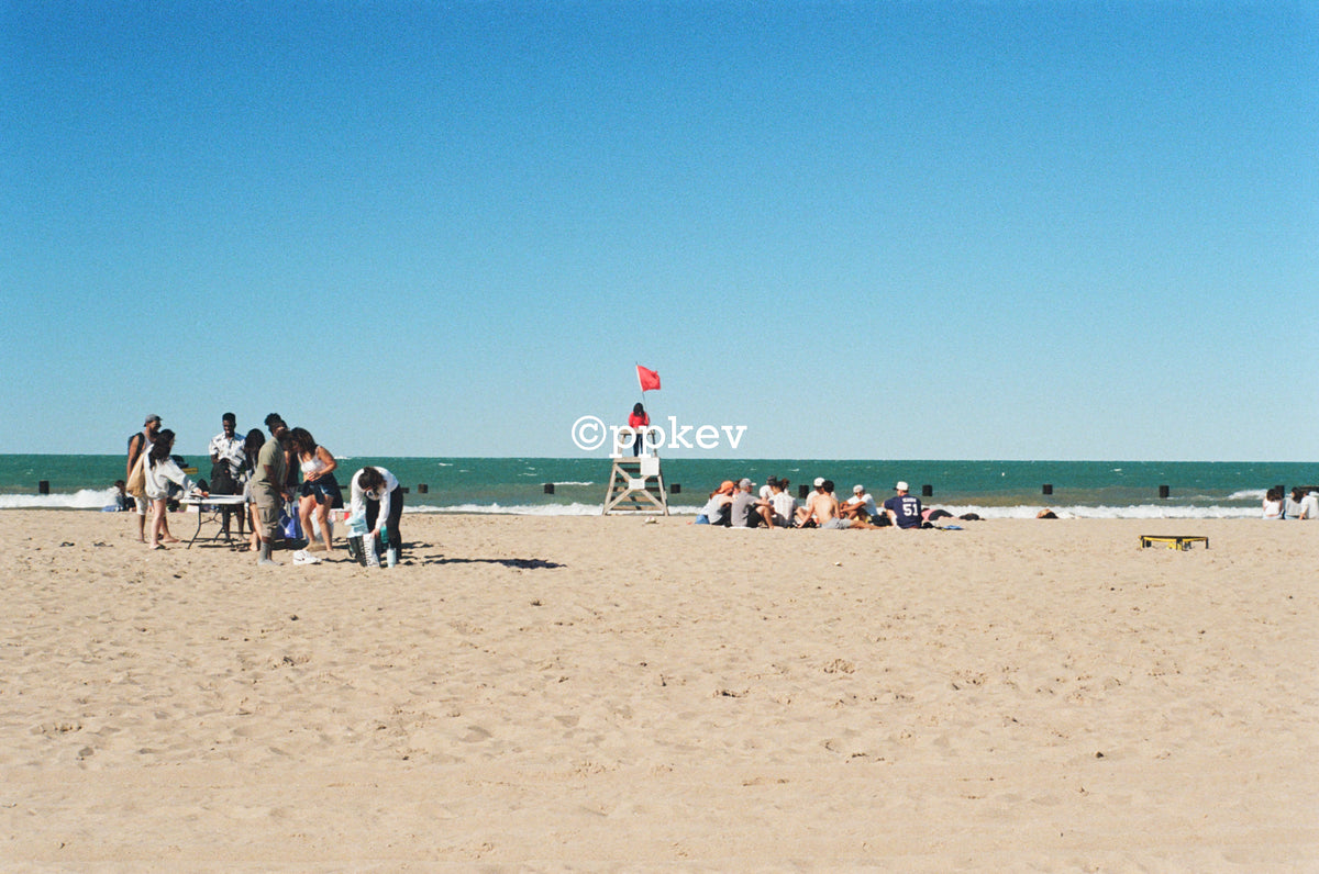 A wide beach scene with sand in the foreground, ocean water in the distance, several small groups of people near the shoreline, and a lifeguard tower with a red flag under a clear blue sky.