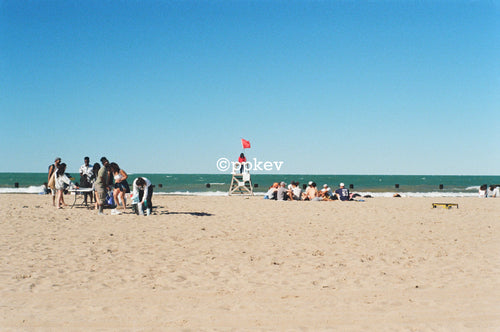 A wide beach scene with sand in the foreground, ocean water in the distance, several small groups of people near the shoreline, and a lifeguard tower with a red flag under a clear blue sky.